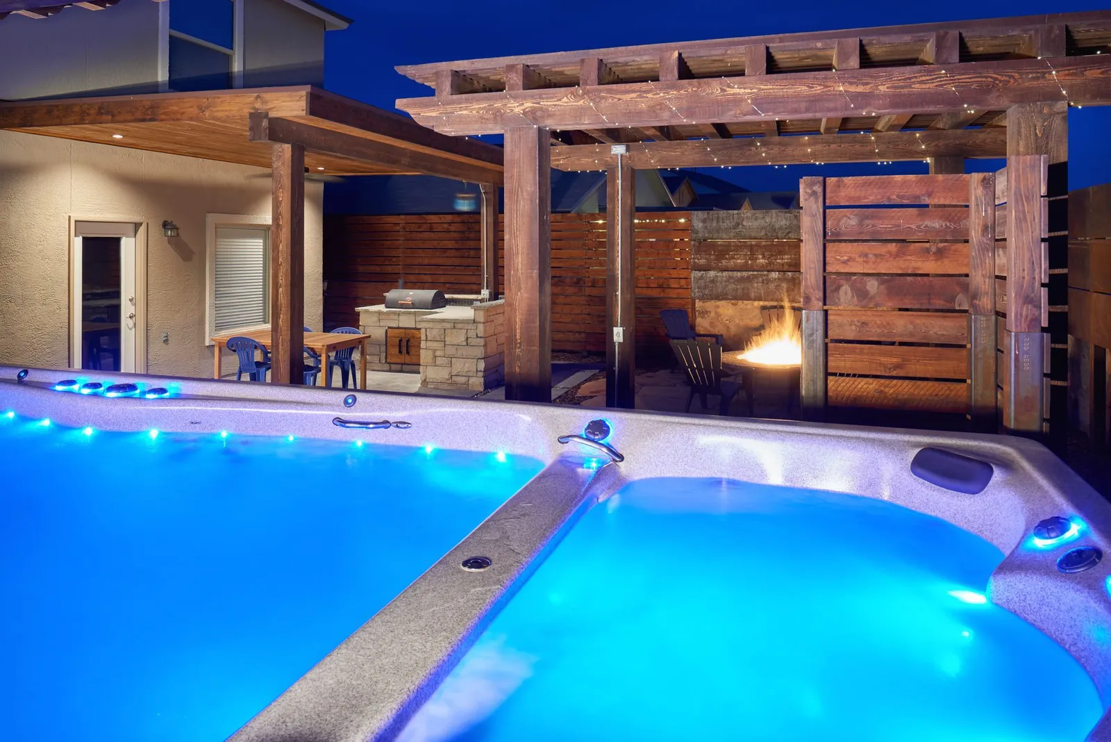 Nighttime pool area with blue illuminated water, a wooden pergola, and a built-in outdoor kitchen.