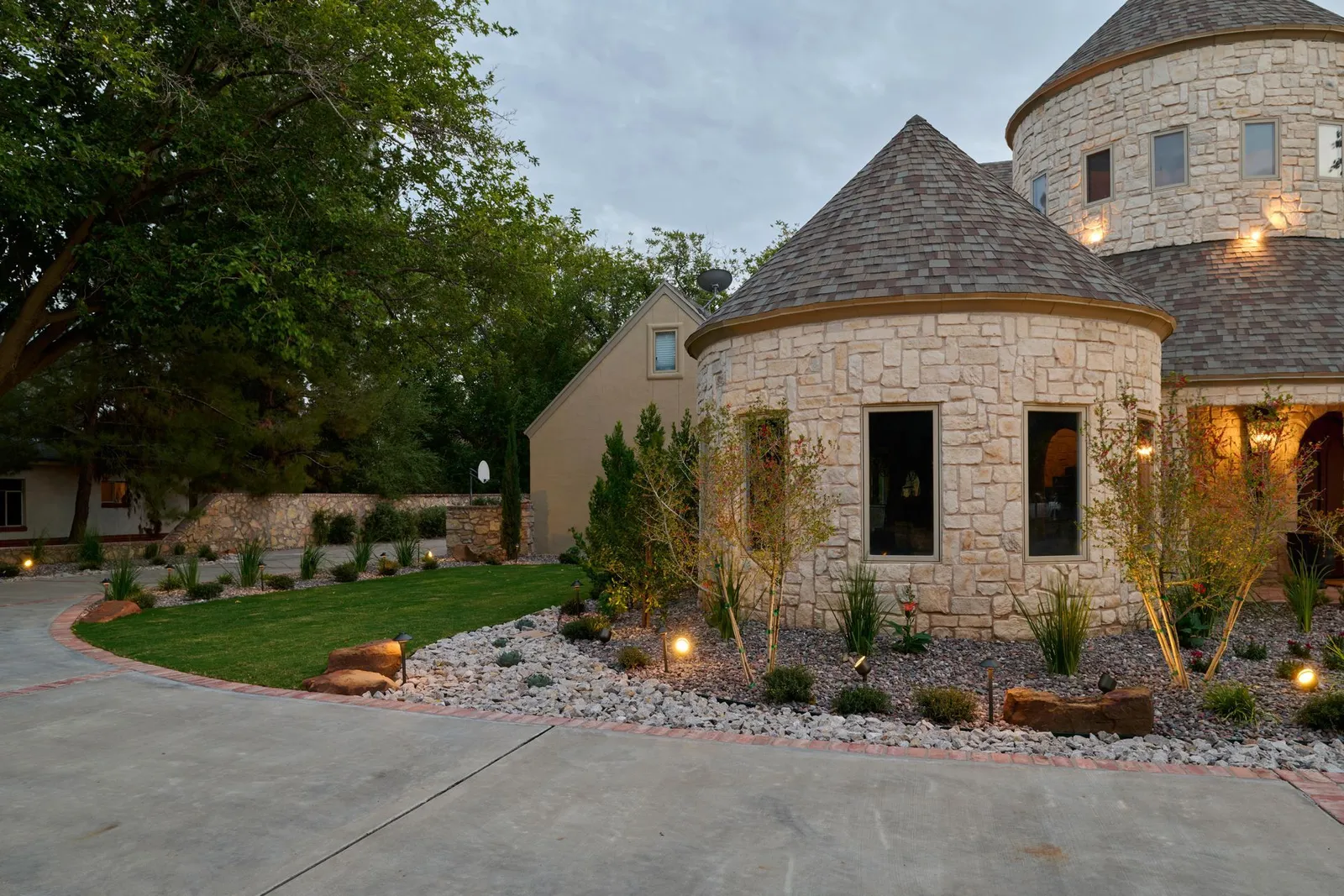 A curving drive and landscape lighting in front of a stone home at dusk.