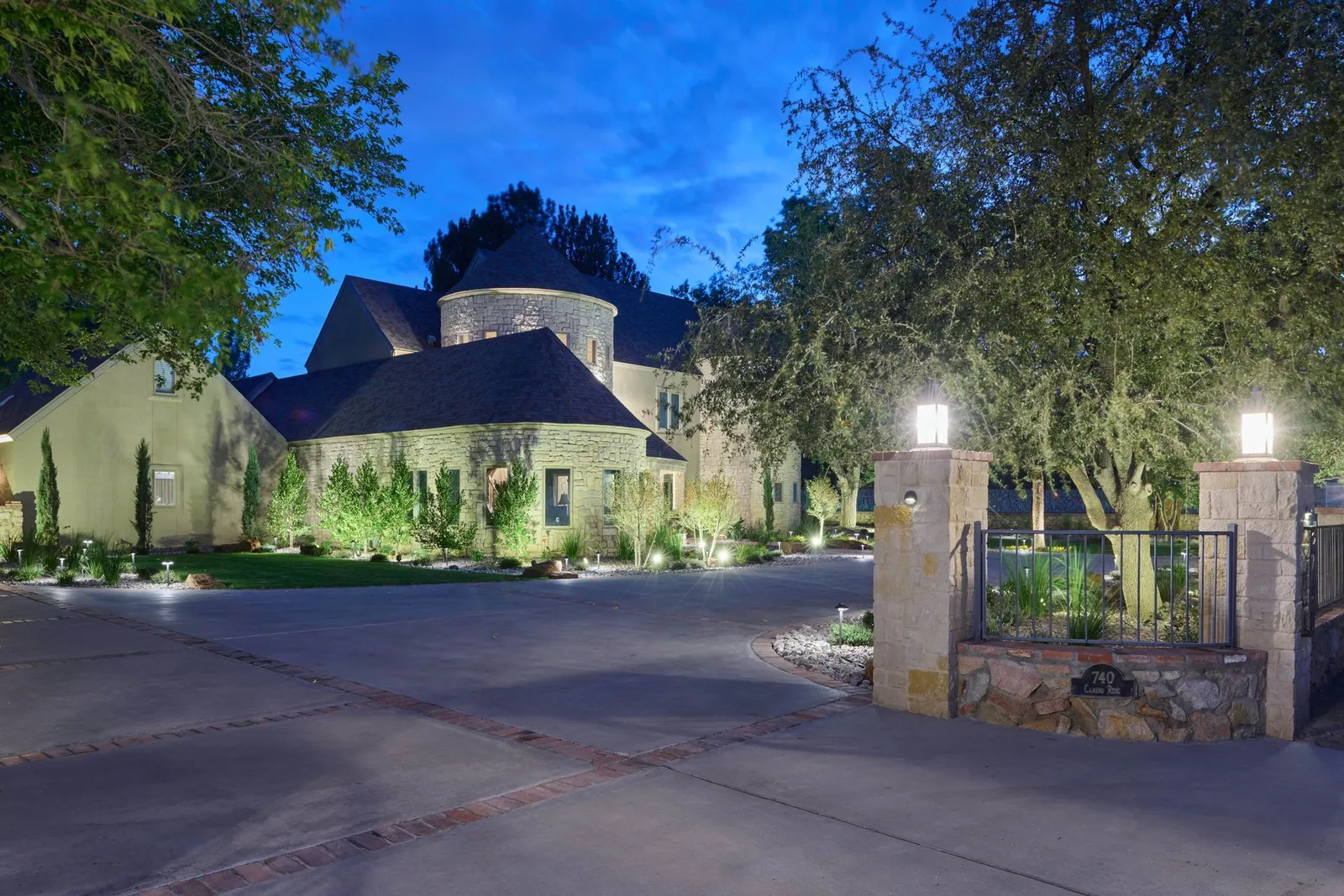 Home at dusk with an illuminated drive, gate, and mature trees.