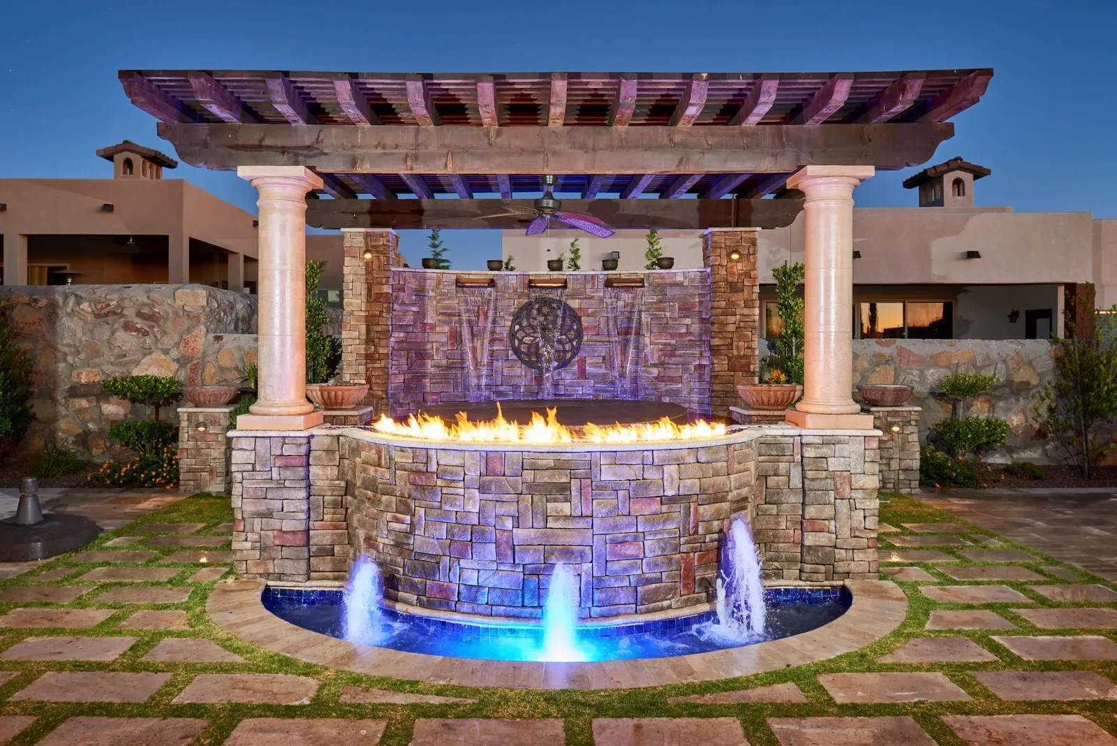 Stone fountain wall beneath a pergola, lit in blue and amber at dusk.