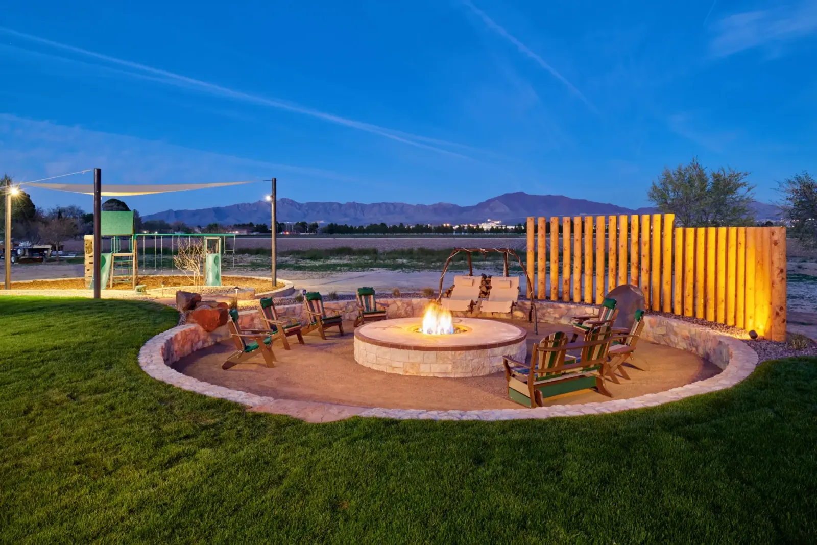 Fire pit area with seating, wooden fence, grass lawn, and mountains at dusk.