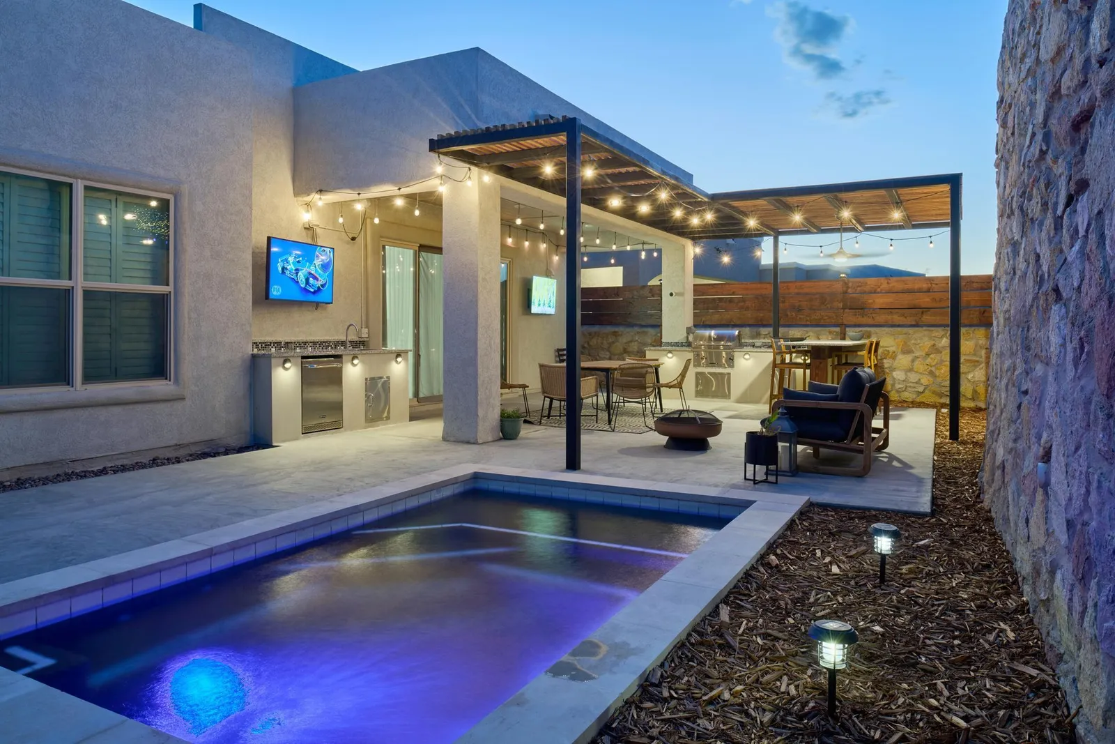 Backyard patio with pool, outdoor kitchen, seating area, string lights, and a pergola at dusk.