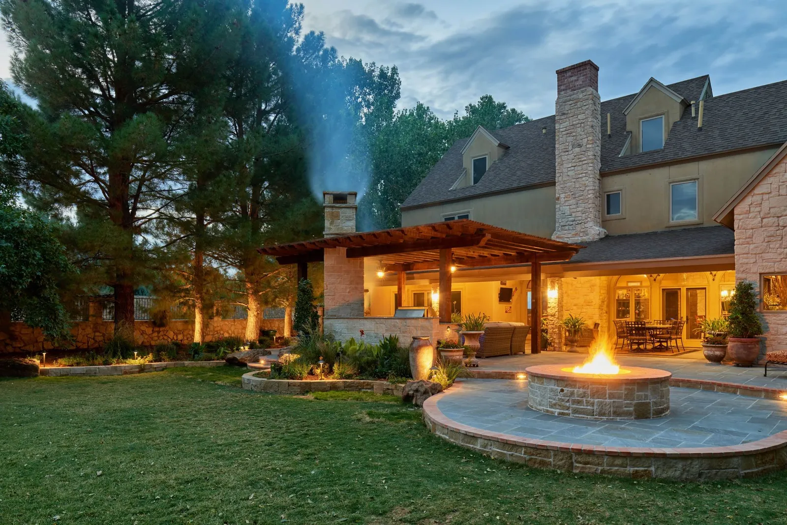 Backyard with fire pit, pergola, and house lit up at dusk.
