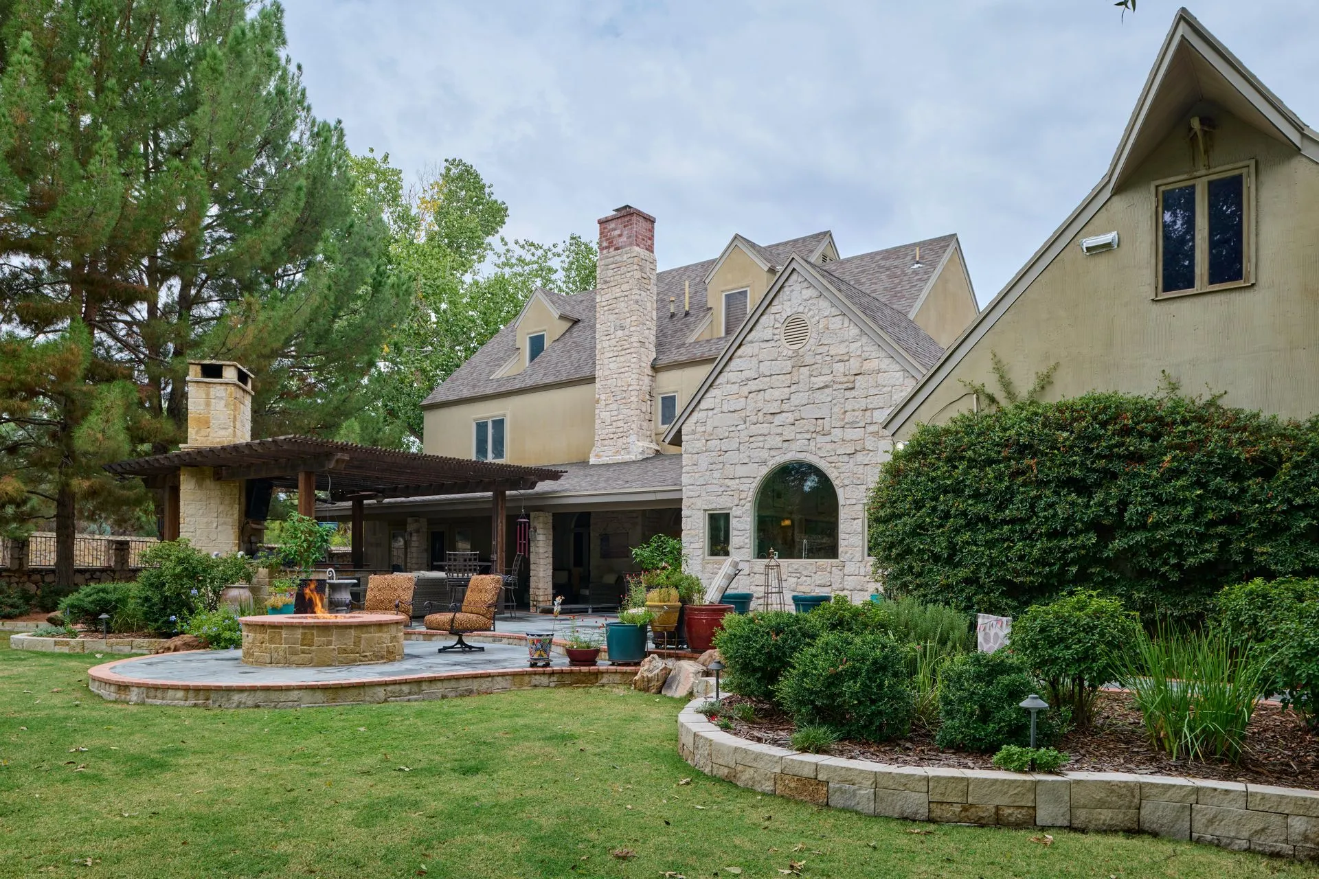 Backyard with house, patio, fire pit, and landscaping under cloudy sky.