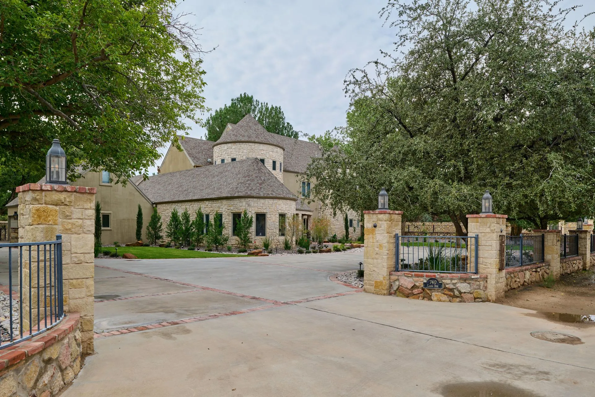 Stone home with a circular driveway and gate posts with lanterns; overcast sky.