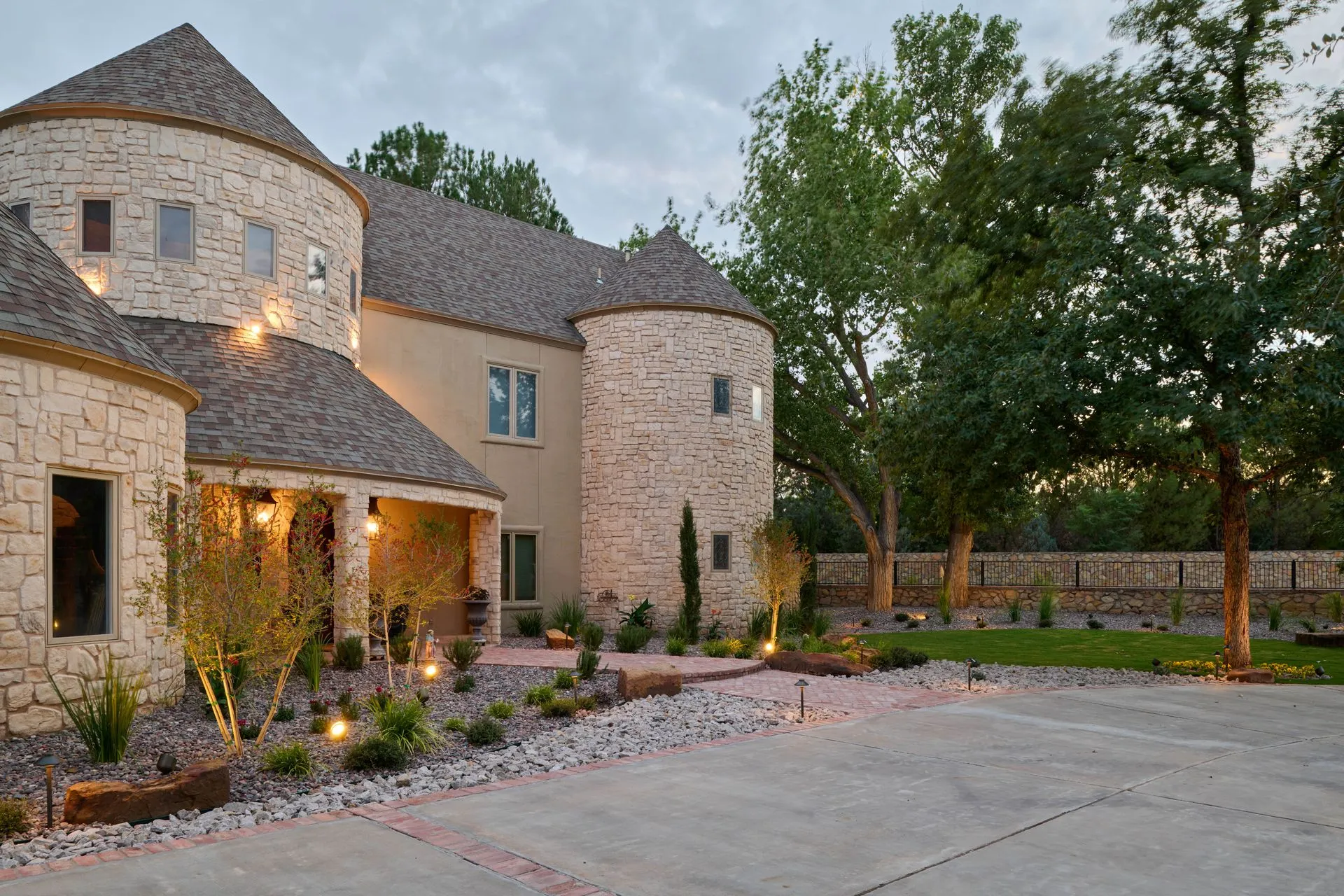 Stone castle-style house with round turrets, concrete driveway, and landscaped yard at dusk.