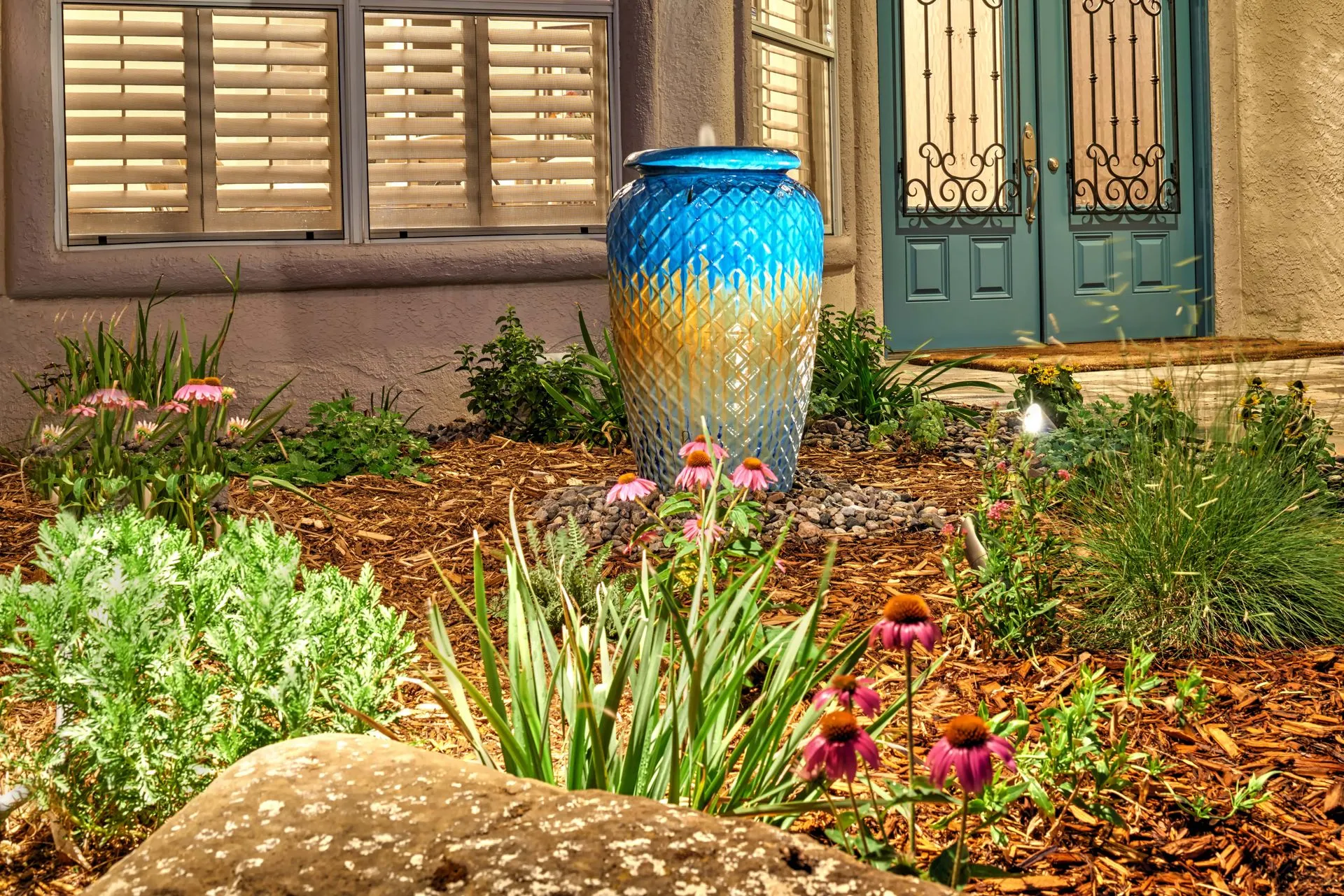 Blue and gold decorative fountain in a garden bed with blooming pink flowers.