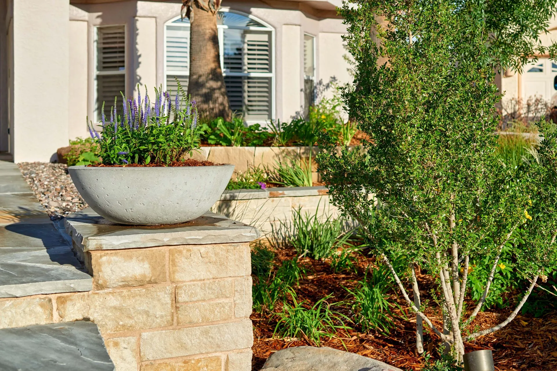 Large stone planter with lavender flowers on a brick pillar, near landscaped garden.