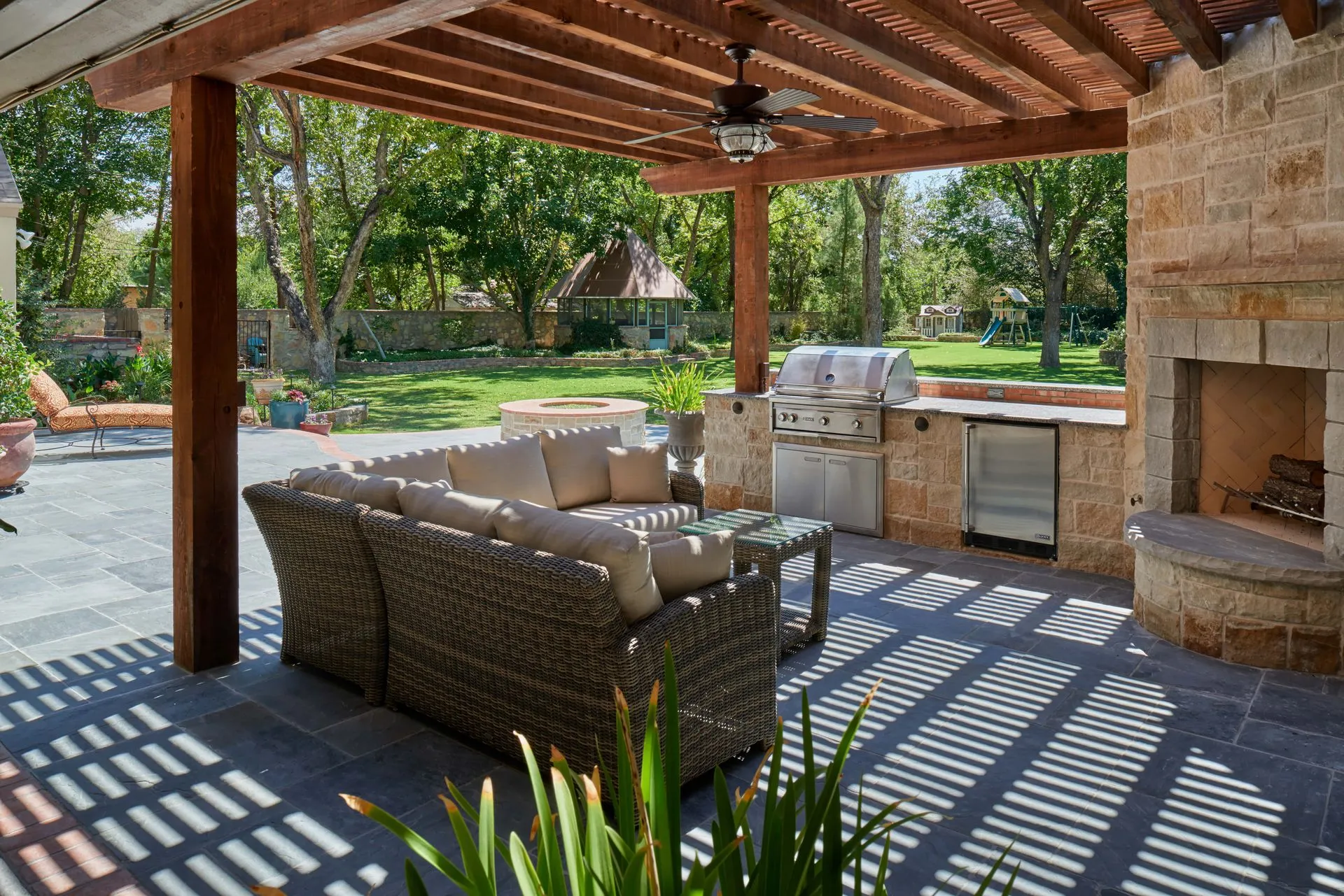 Outdoor patio with sectional sofa, grill, fireplace, and pergola, in a backyard setting.
