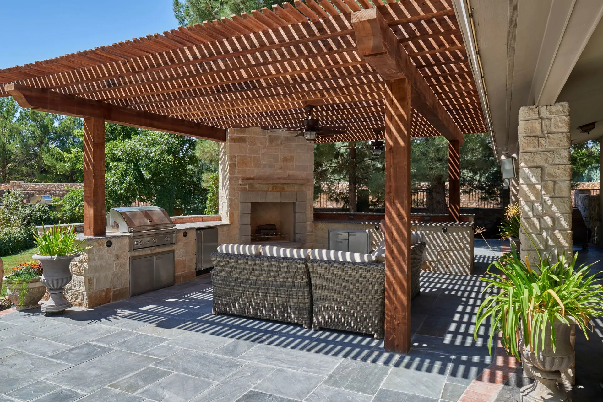 Outdoor patio with pergola, fireplace, grill, and seating. Sunlight casts shadows on the stone and dark pavers.