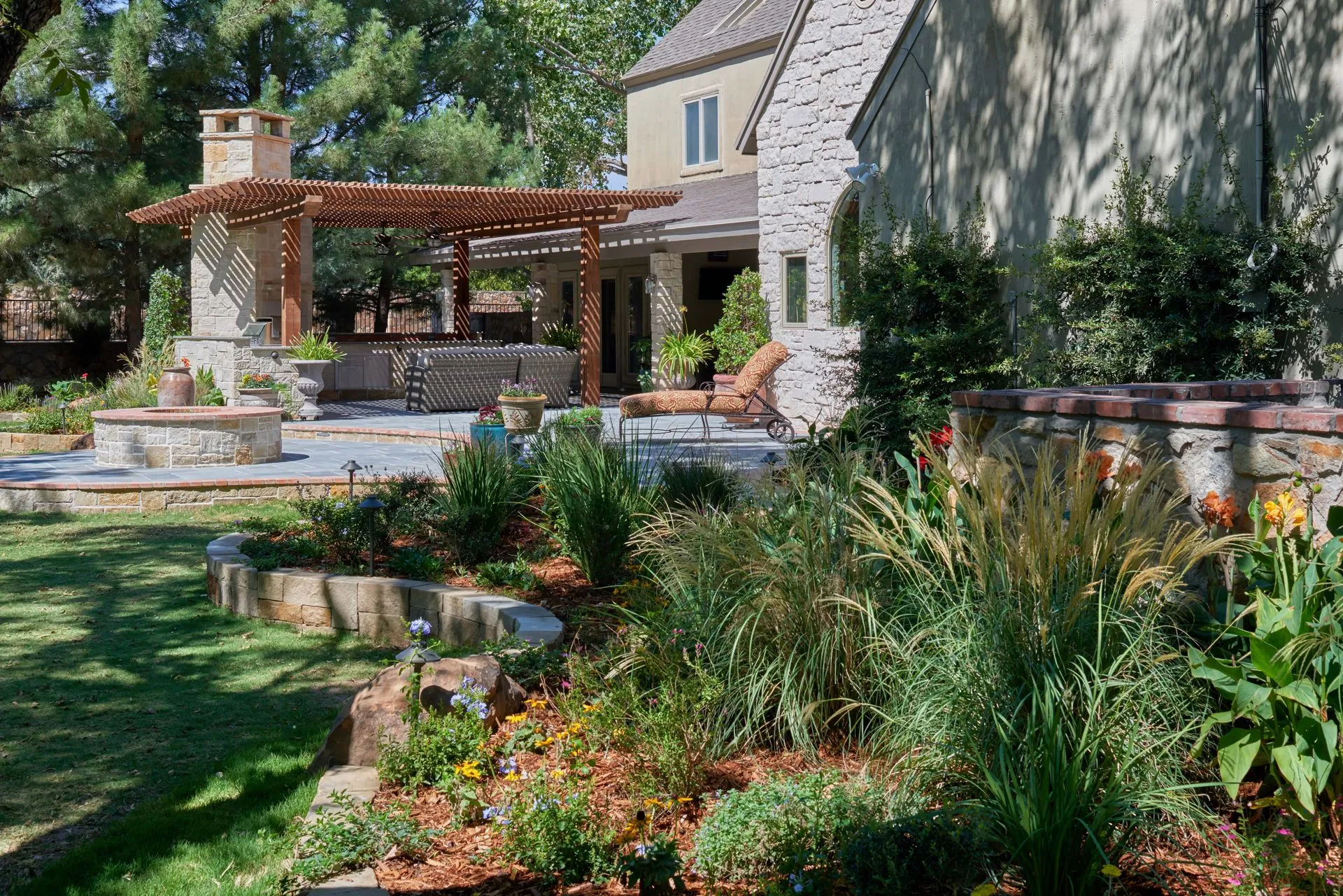 Stone patio and pergola with fireplace, lush landscaping, and a house in the background.