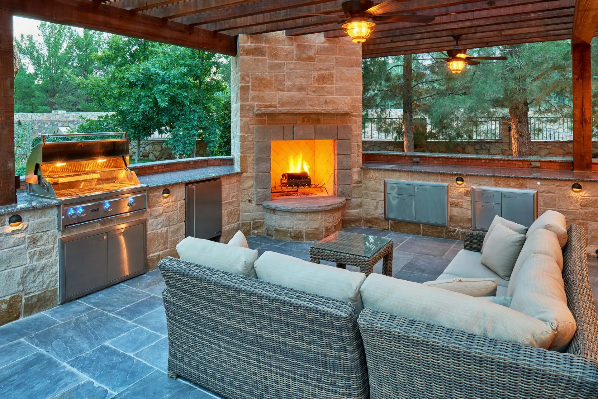 Outdoor kitchen with stone walls, fireplace, grill, and wicker seating under a wooden pergola.