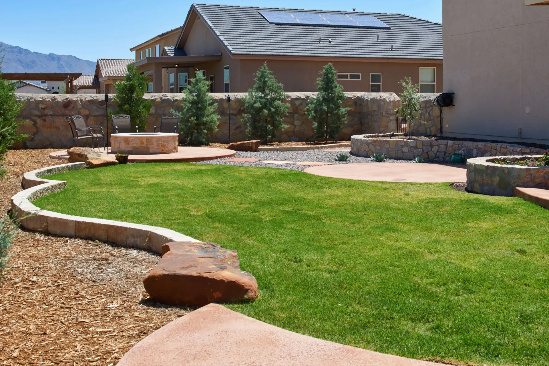 Backyard with a green lawn, stone patio, fire pit, and surrounding landscaping.