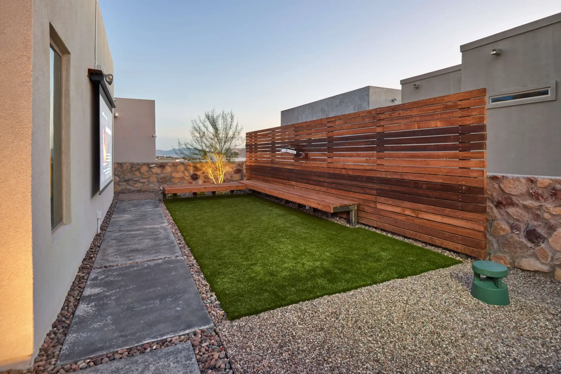 Side yard with artificial turf, a wood privacy screen, bench seating, gravel, and surrounding stucco homes.