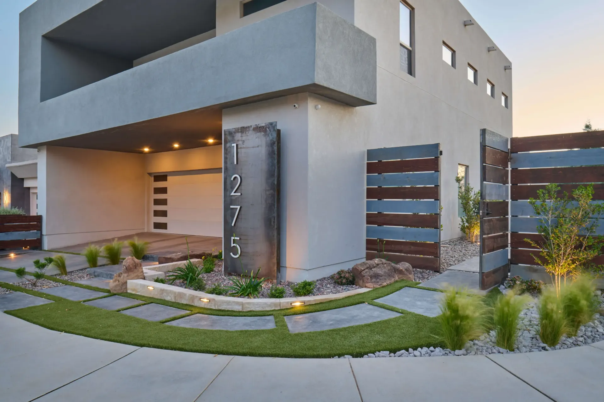 Modern home exterior with gray stucco, wood accents, a landscaped front yard, and an address marker.