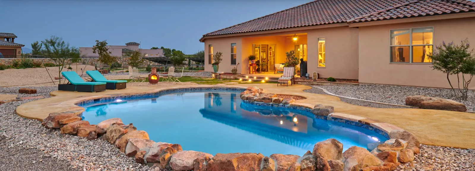 Pool with turquoise water surrounded by rocks and a house with lights on in the background.