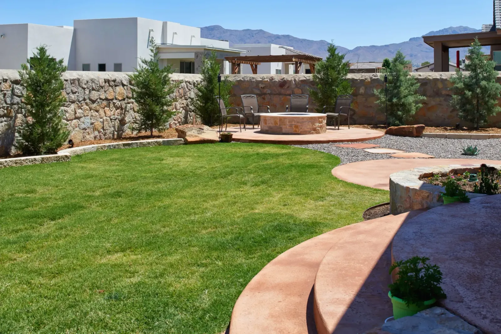 A lush green backyard with a fire pit, bordered by a stone wall and trees, under a clear blue sky.