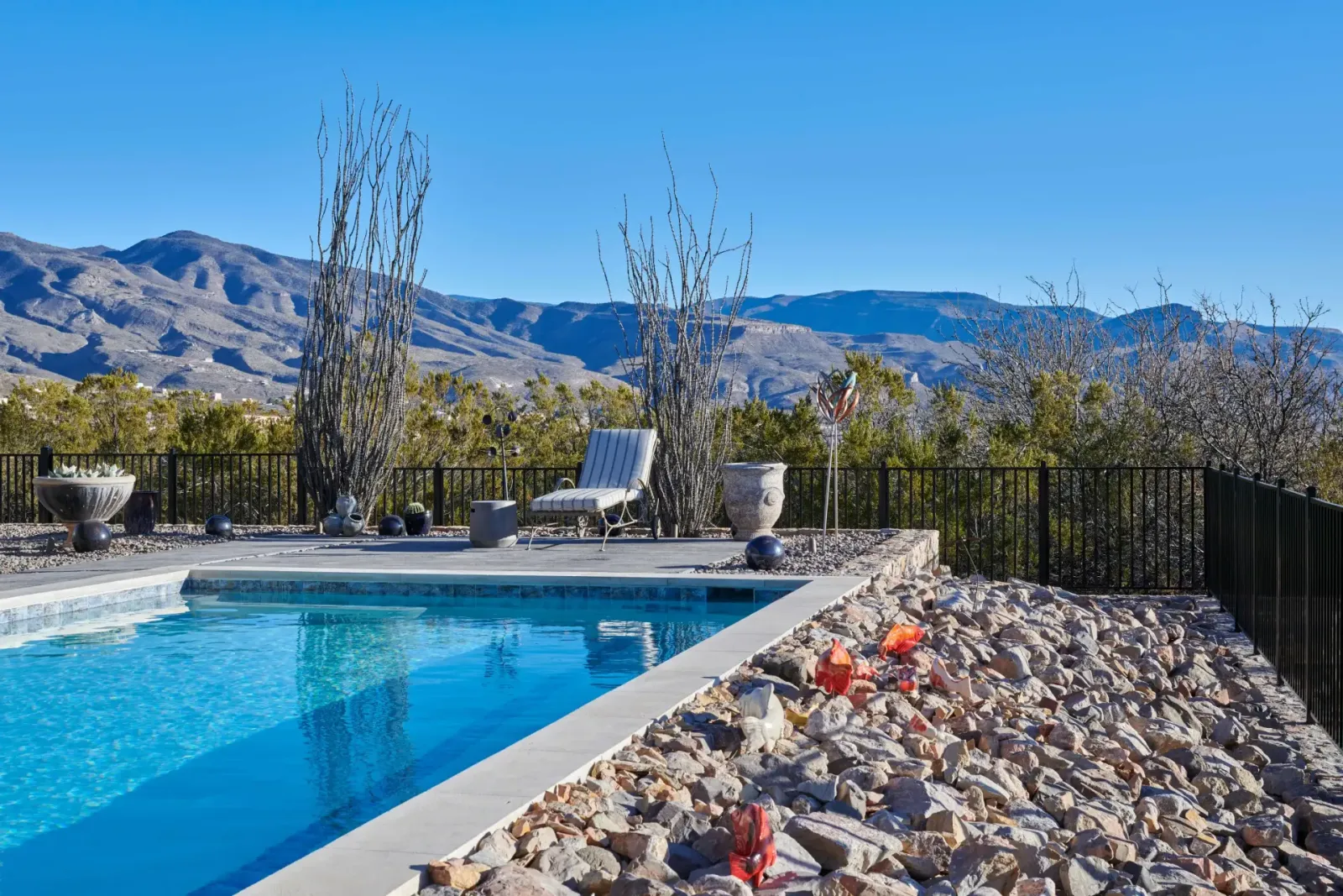 Pool with mountain view, blue water, beige rocks, black fence, and white chair.