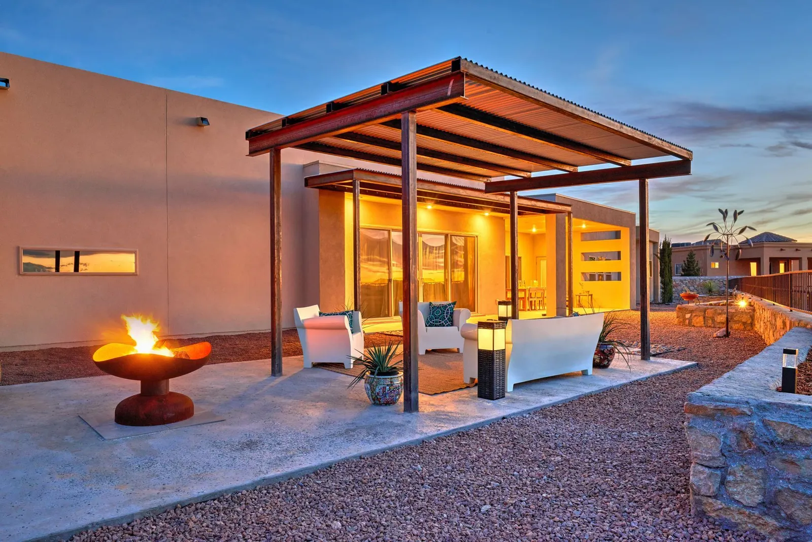 Covered patio with seating and a fire pit at dusk, with warm lighting.
