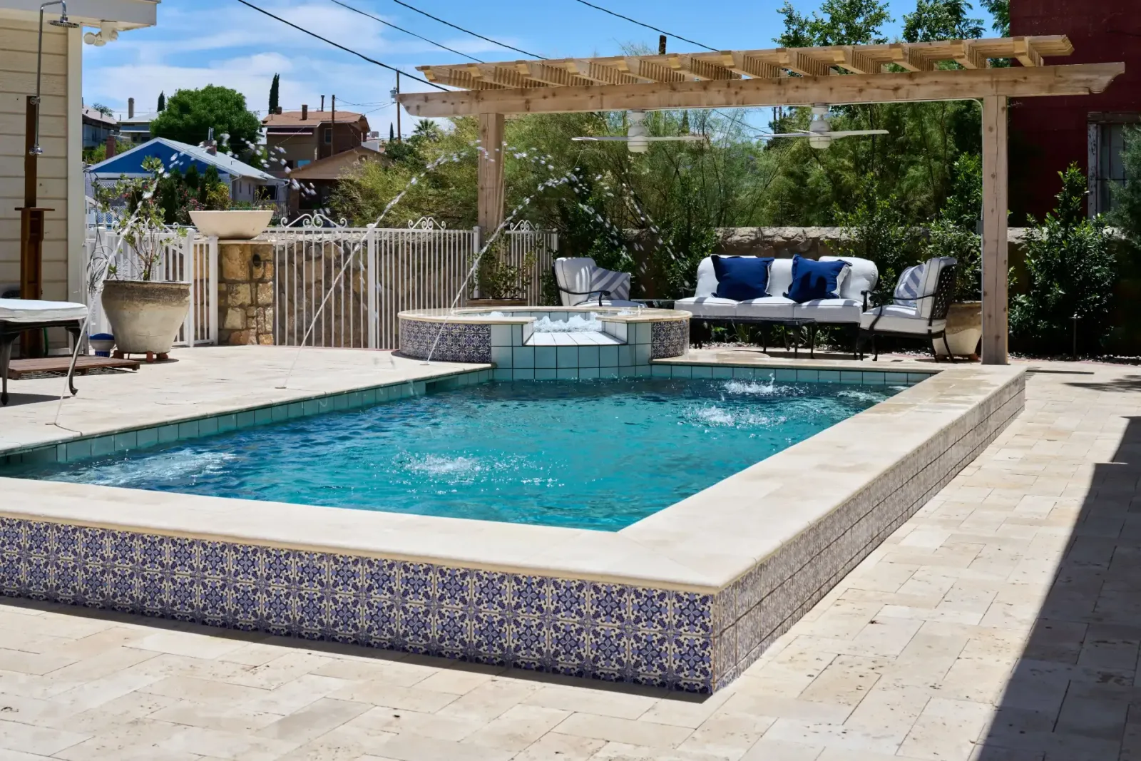 Outdoor pool with blue mosaic tile, travertine patio, and lounge area under a pergola.