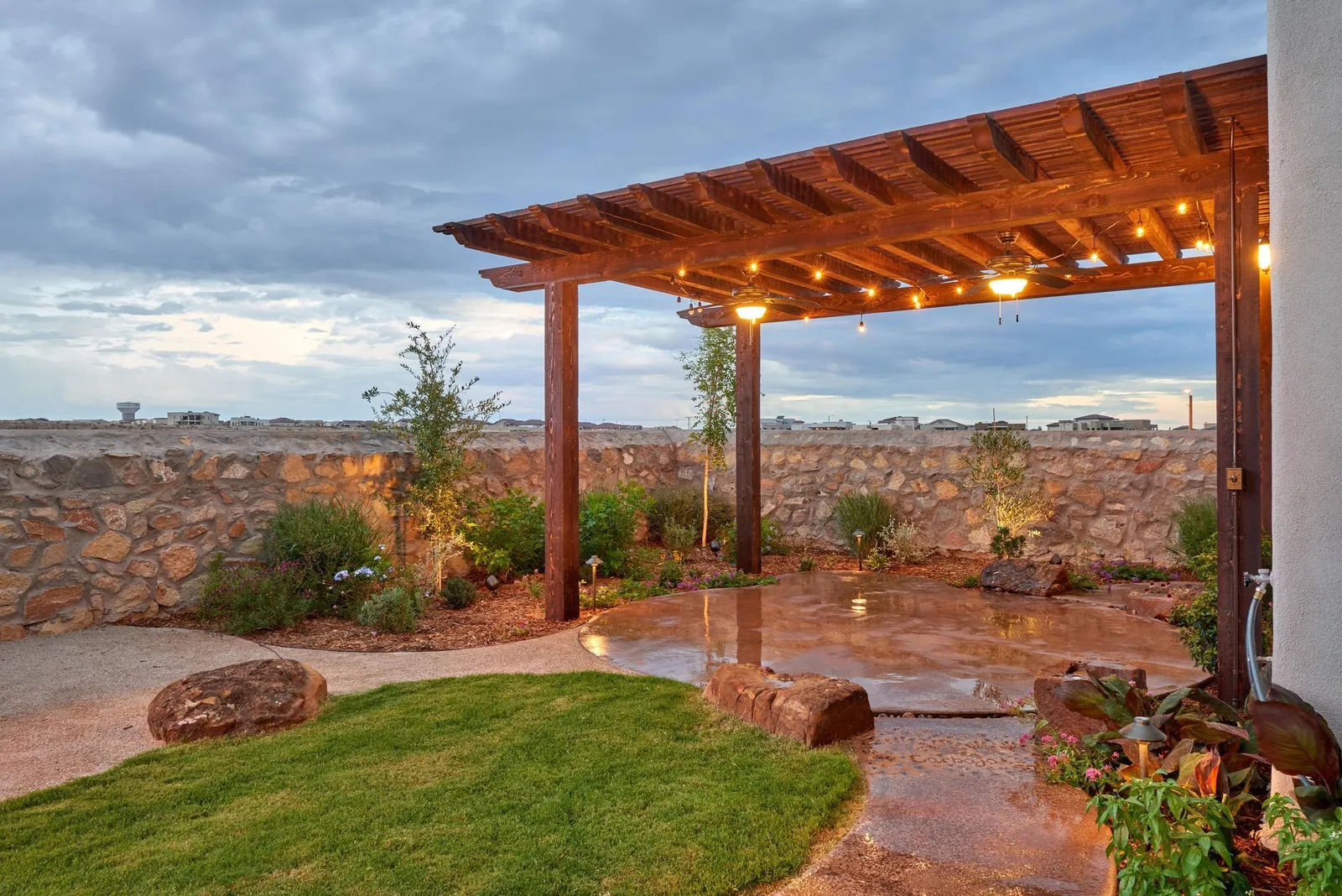 Patio with wooden pergola, string lights, and small garden area, stone wall background. Overcast sky.