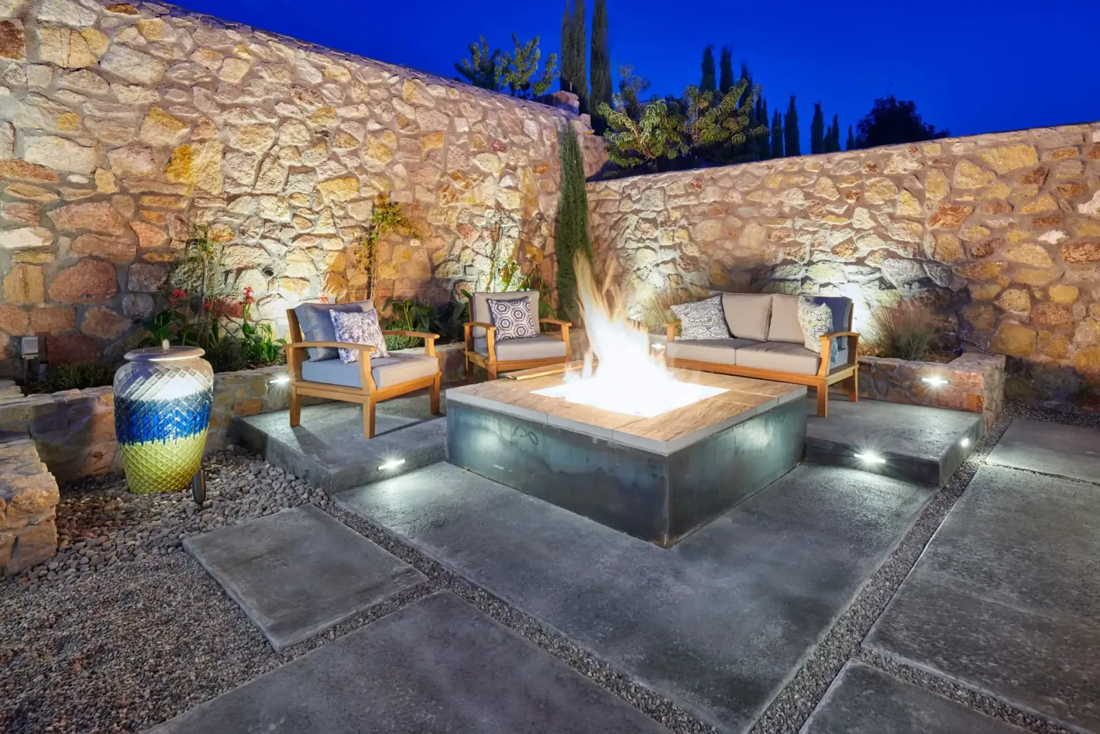 Outdoor stone patio with seating around a lit fire pit at dusk.
