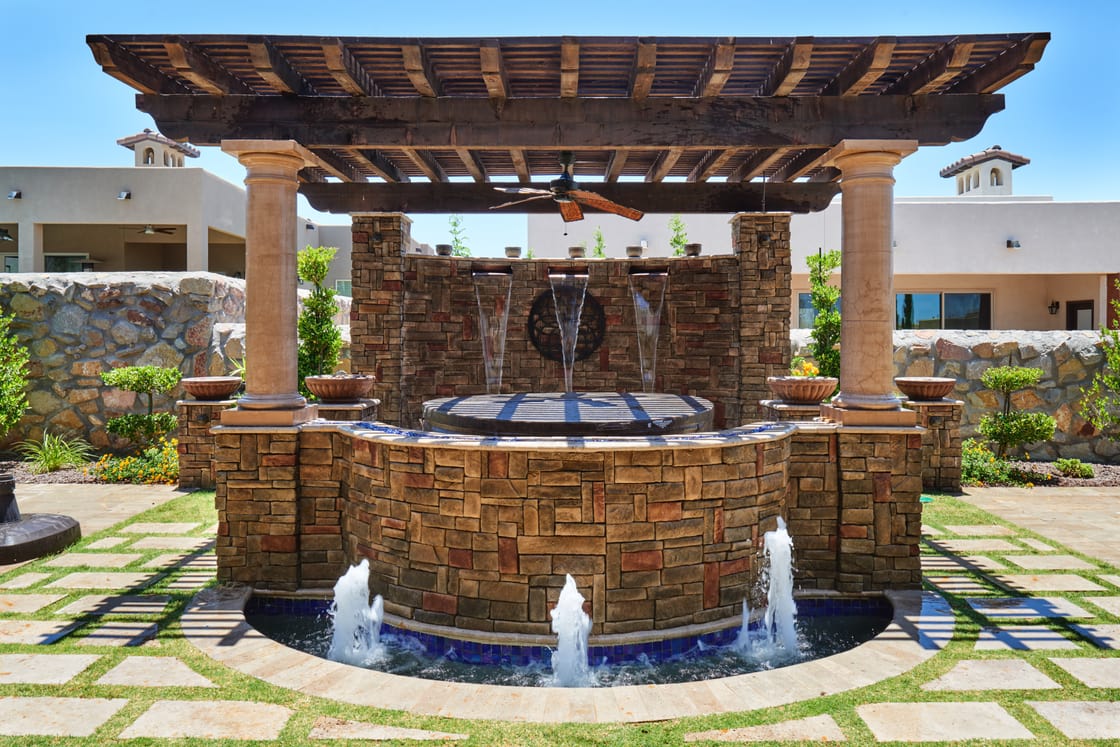 Water jets around a round stone fountain beneath a timber pergola.