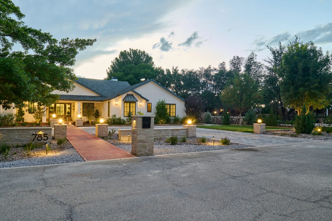 Low lights illuminate a planted residential entrance and broad paved drive at dusk.