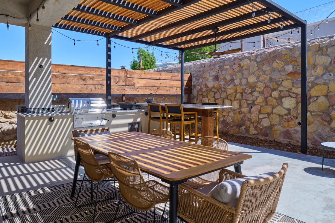 An outdoor kitchen and dining table under a slatted pergola beside a stone wall.
