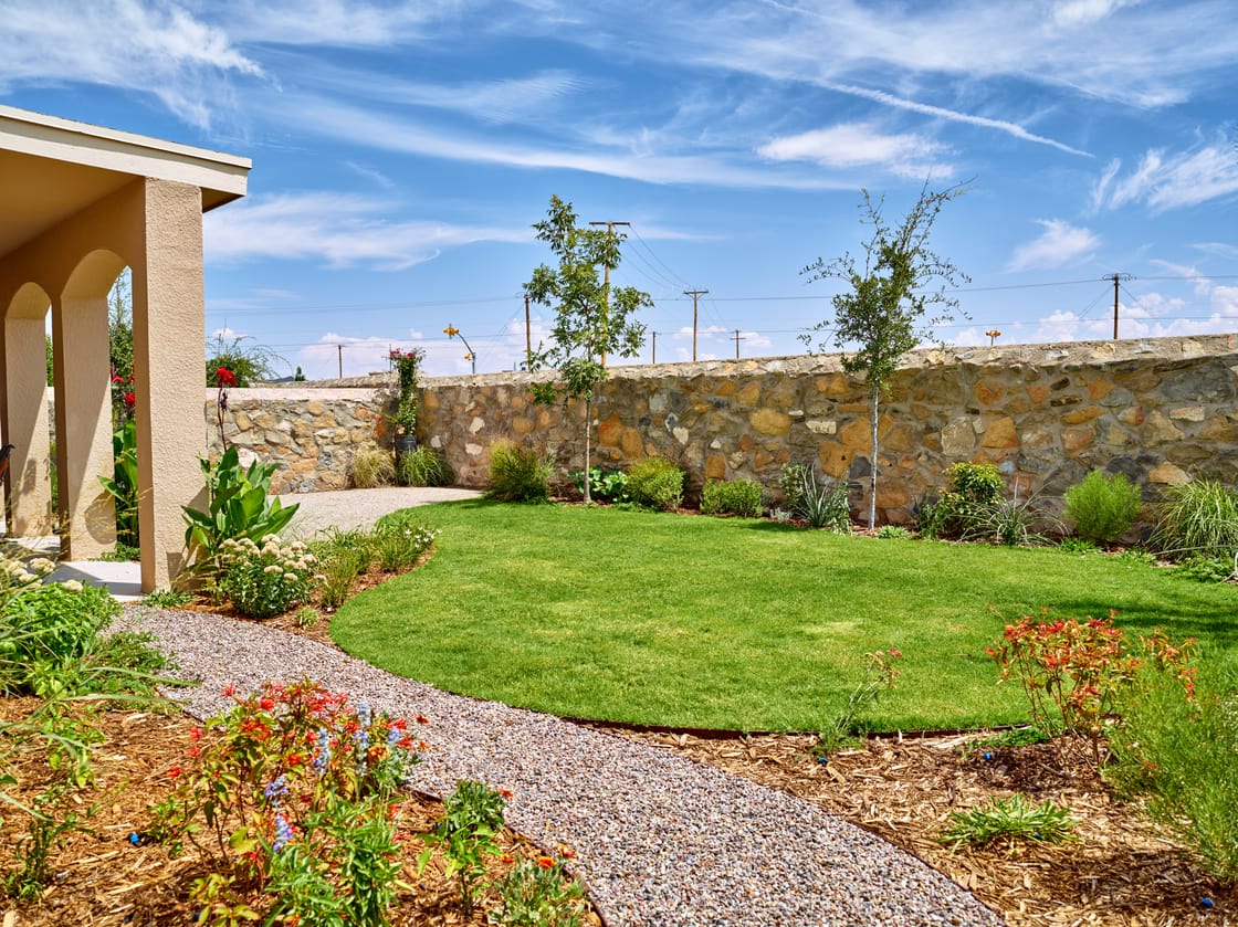A curved lawn bordered by flowering plants, gravel, and a stone wall.