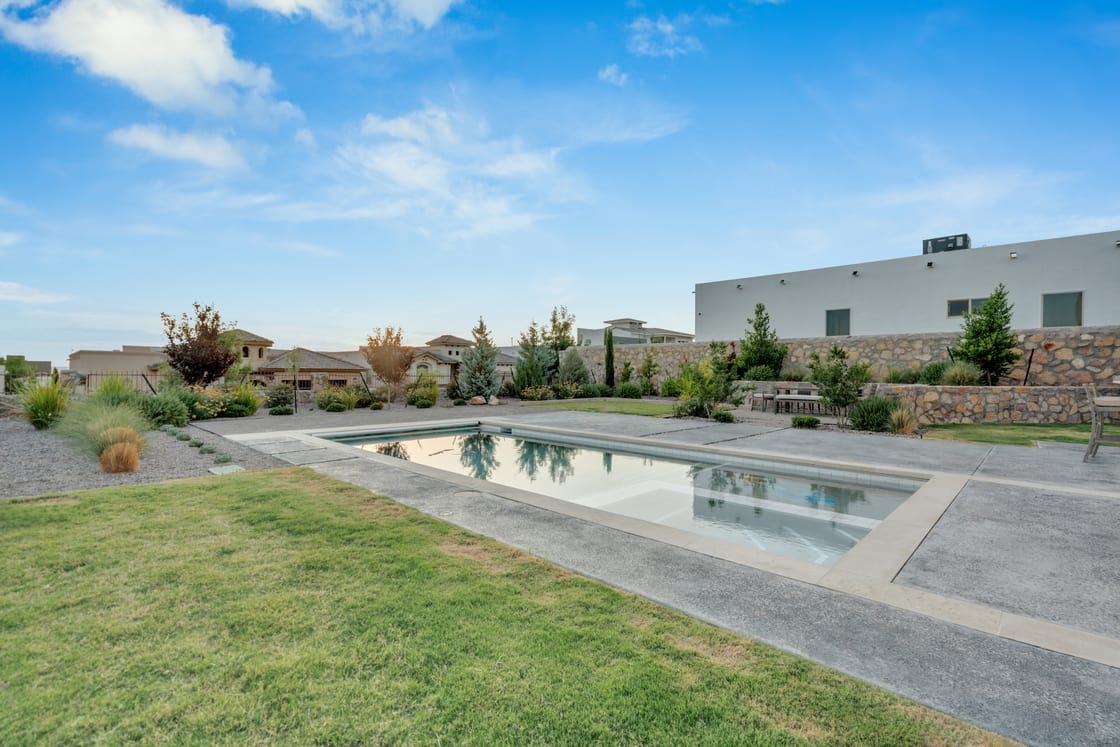 A rectangular pool with pale coping beside a lawn, patio, and planted stone boundary.