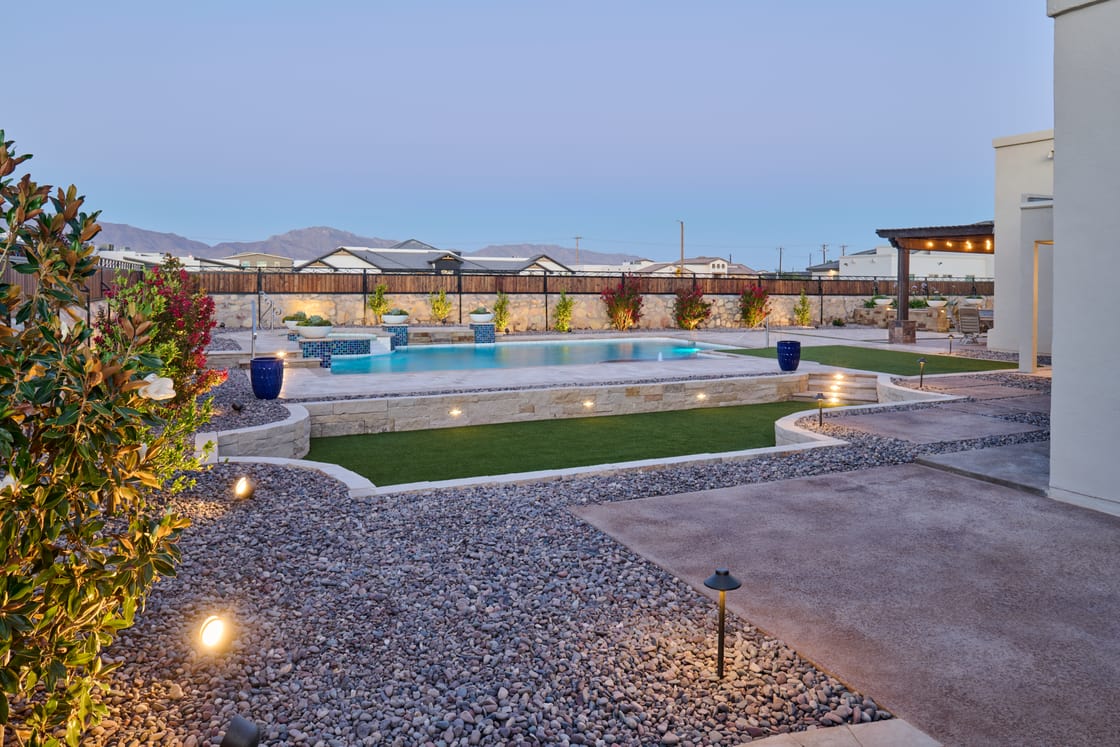 A backyard pool with planting and a basketball hoop beyond the water at dusk.