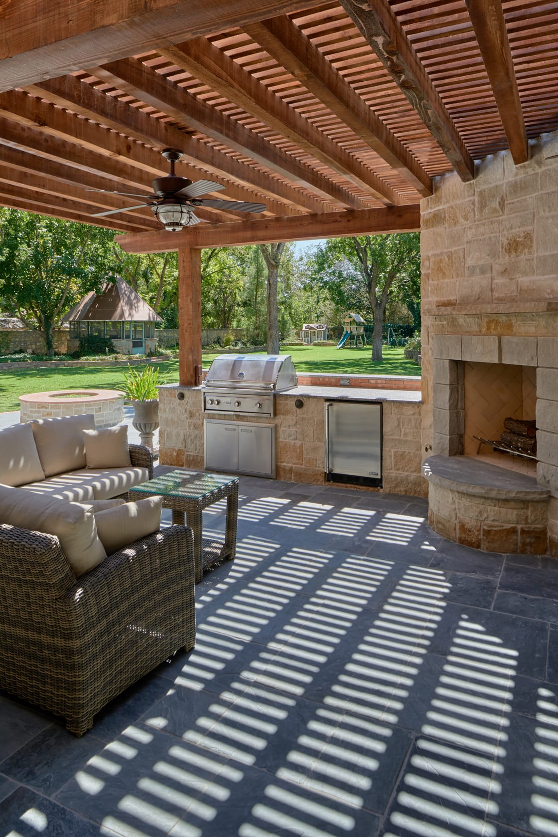 A grill, stone fireplace, and seating beneath a pergola casting striped shadows.